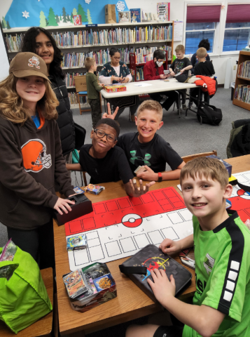 A group of kids and librarians sit around two tables playing with pokémon cards