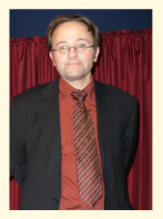 Photo of a man in a red shirt and tie and a black jacket in front of a red background.