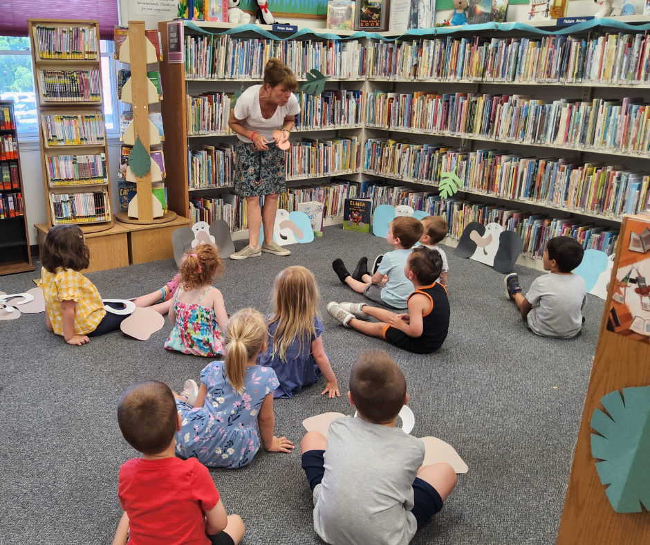 A group of children sit on the floor while a librarian guides them in an activity.